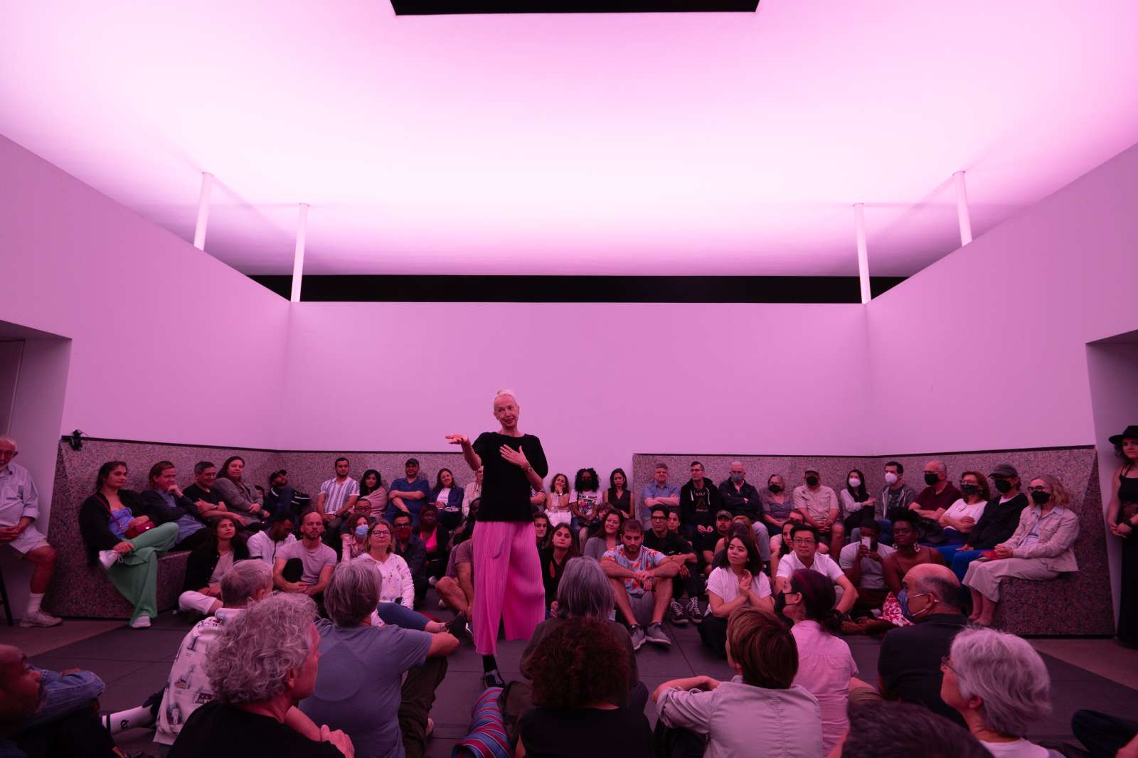 Karole Armitage speaking to an audience in the skyspace wearing a black shirt and pink pants.