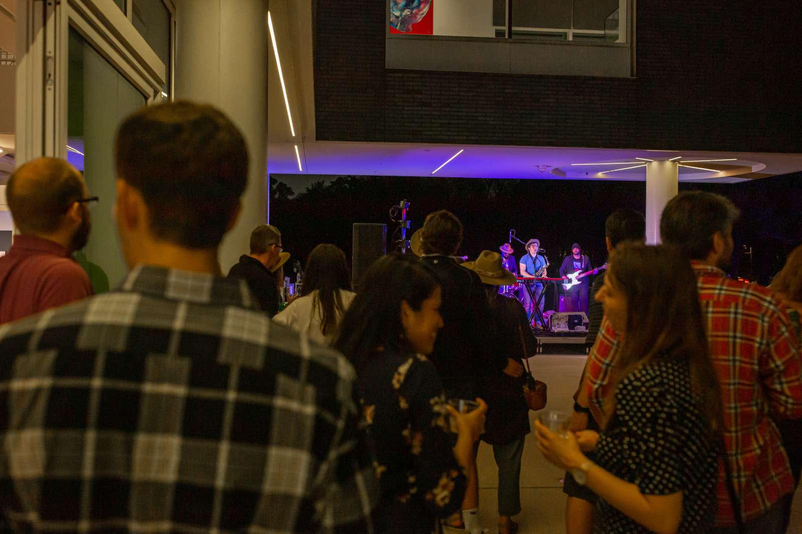 In the foreground, audience members stand and listen to performer who is in the background